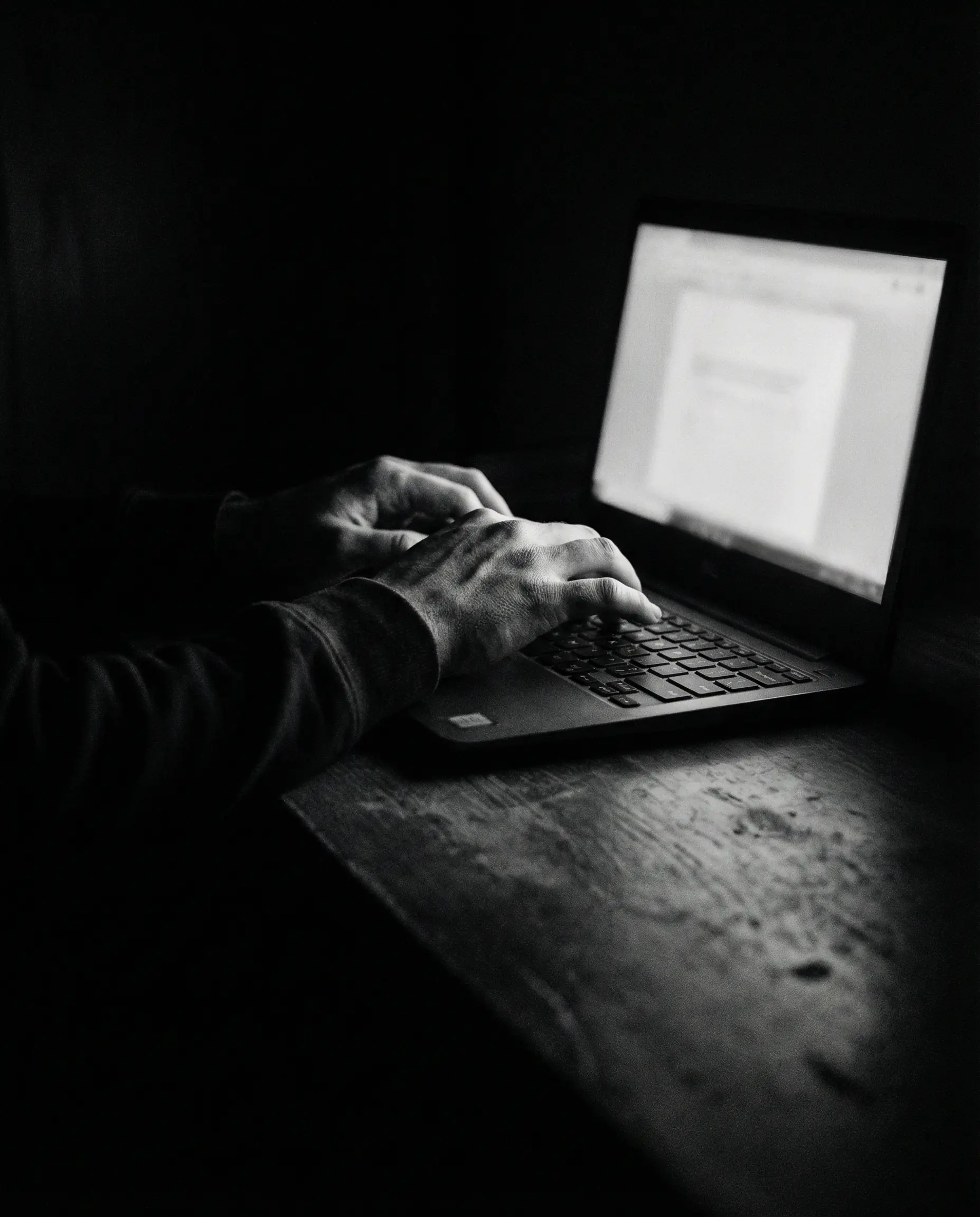 Hands typing on laptop in dark room with screen glow